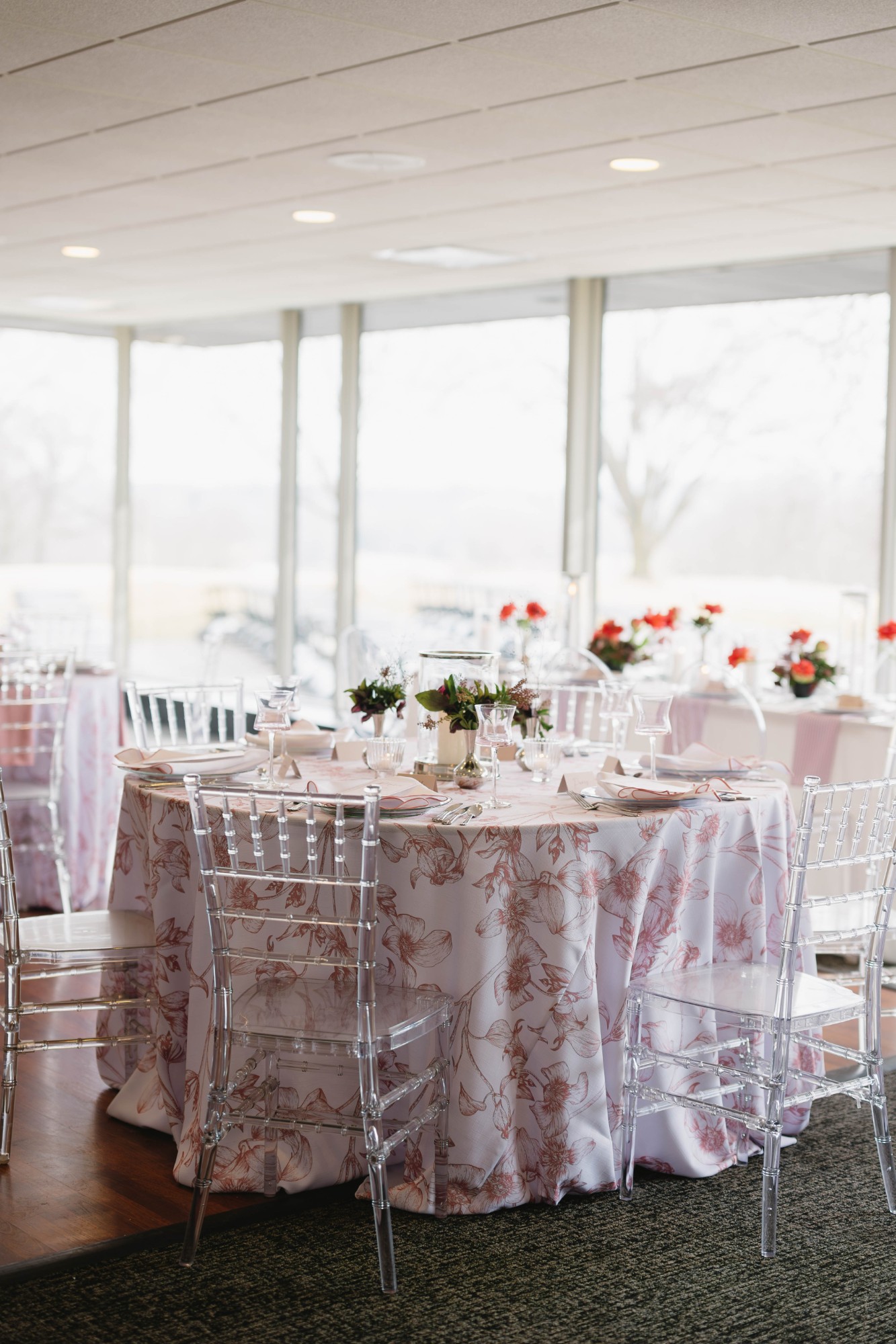 A round table set with blush floral-toile linen and clear acrylic chiavari chairs in a glass-walled room, with red rose centerpieces on tables beyond
