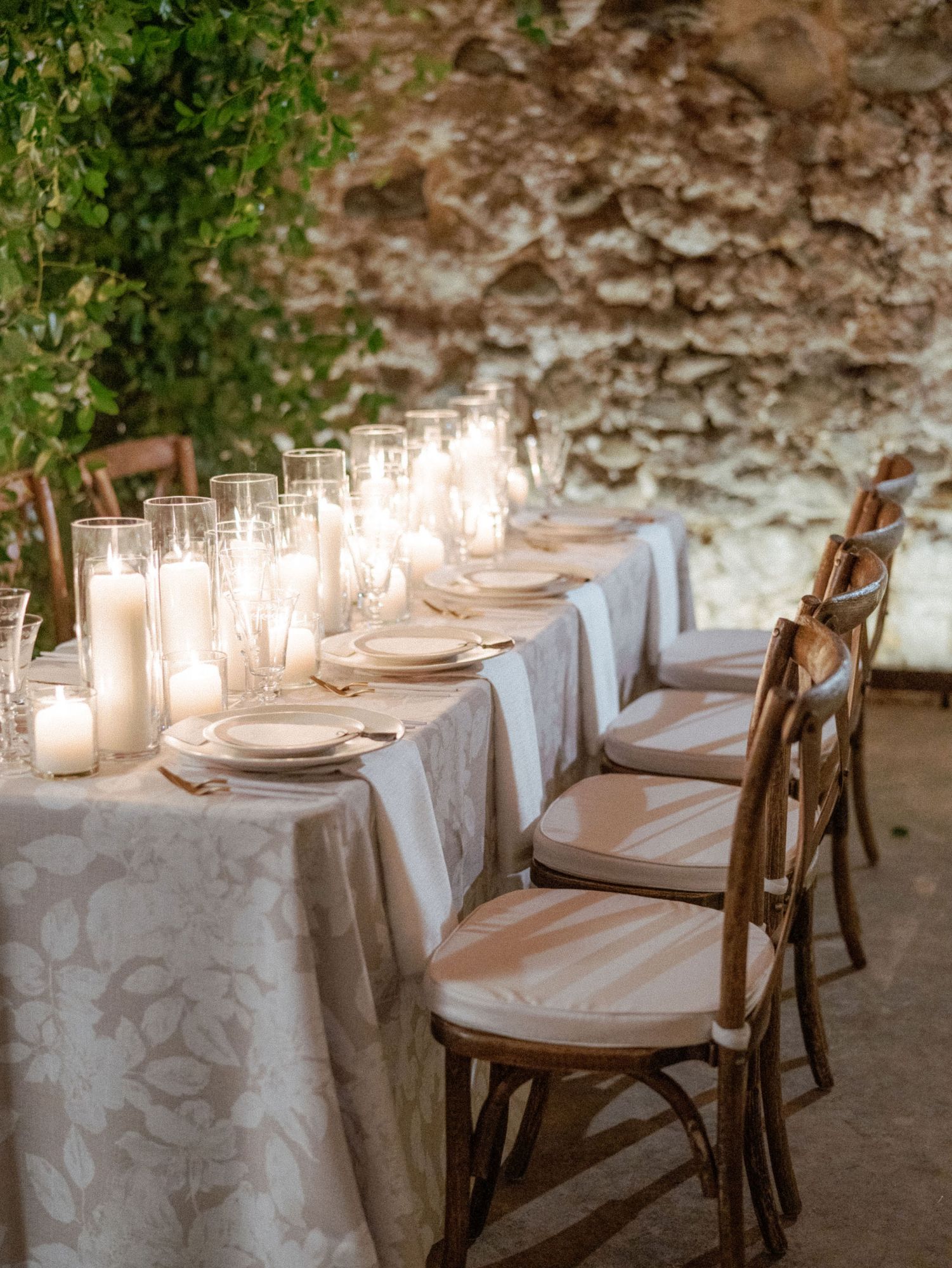 A long candlelit table against a rustic stone cellar wall, set with cream floral-damask linen, tall glass hurricane candles, pillar candles and gold flatware, with cross-back wood chairs and trailing greenery overhead